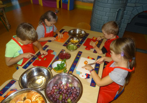 Grupa dzieci siedzi przy stole, w ręku trzymają noże, którymi kroją owoce na desce.